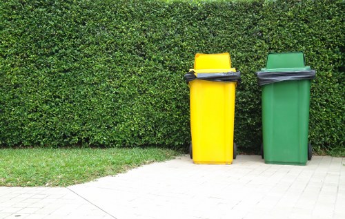Two large wheelie bins, one yellow and one green, positioned side by side on a paved surface in front of a neatly trimmed green hedge. The yellow bin, located to the left, has a closed lid with a black handle and is filled with dark waste material, while the green bin on the right, also with a closed lid, appears to contain similar waste. The bins are made of durable plastic with textured surfaces, typical for rubbish collection, and are situated outdoors in a residential area. The paved ground and hedge provide a tidy, organized environment, suggesting a typical rubbish disposal area possibly in a suburb, such as Parsons Green or nearby in London, associated with services like those offered by Office Clearance Parsons Green for waste removal and clearance tasks.