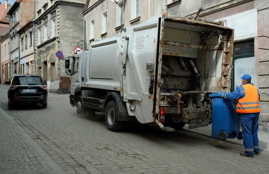 A nighttime scene showing three large blue dumpster bins filled with mixed waste, including black and clear plastic bags, cardboard boxes, and loose rubbish such as paper, plastic, and packaging mater