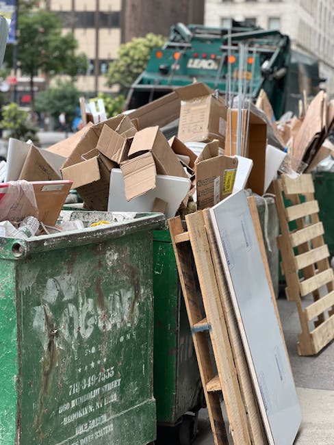 Two large black plastic bags filled with waste, placed on the pavement near the edge of a street in front of a black picket fence. The bags appear to contain household or garden rubbish, with some par