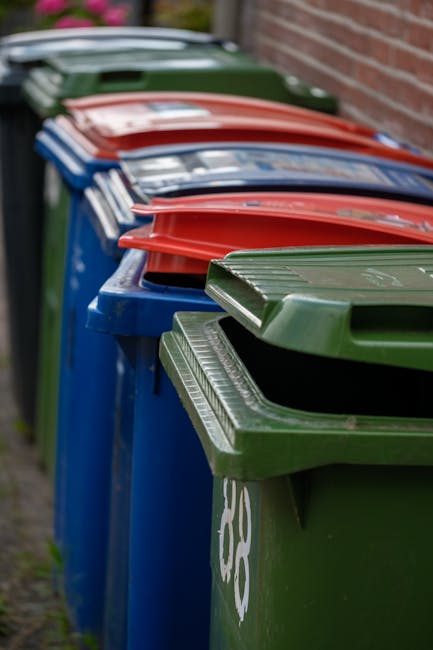 A large green waste disposal skip filled with a variety of packaging materials, including flattened cardboard boxes, paper cartons, and wooden pallets, situated outdoors on a city street. The cardboar