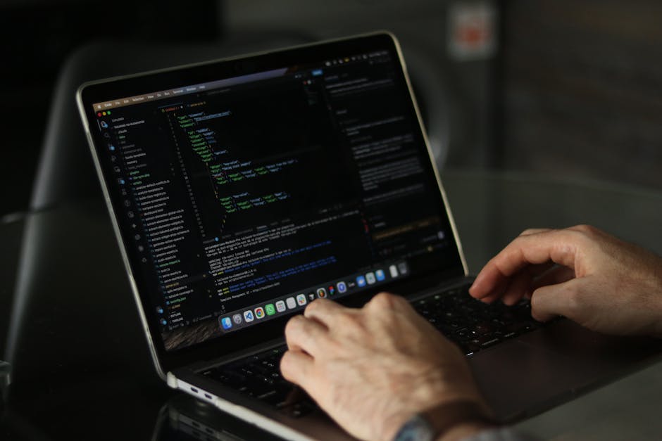 A person using a laptop computer placed on a white surface with a clean, light-colored background. The laptop’s screen displays a code editor with multiple open files, lines of coding, and folder na